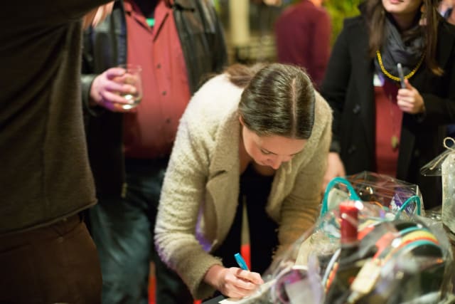 Woman signing a bid sheet at the Love Bomb 2017 charity event for My Voice Music, surrounded by attendees and display items.